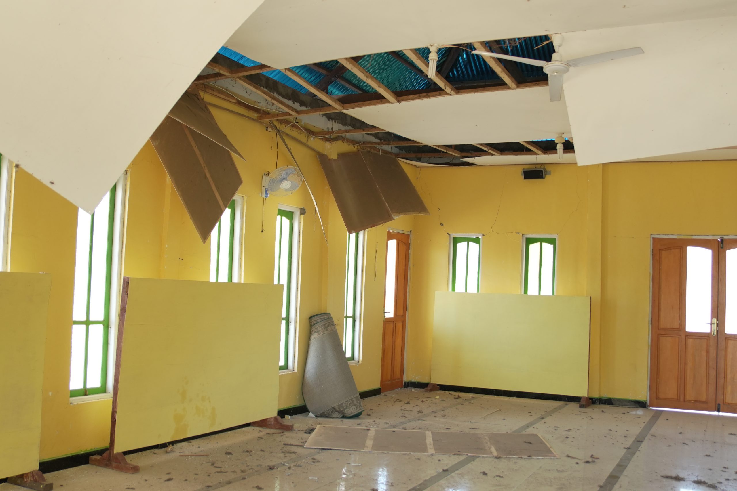 Kondisi interior Masjid Al Muhajirin Waekokak yang rusak parah akibat gempa: plafon runtuh, atap bocor, dan lantai kotor oleh puing.