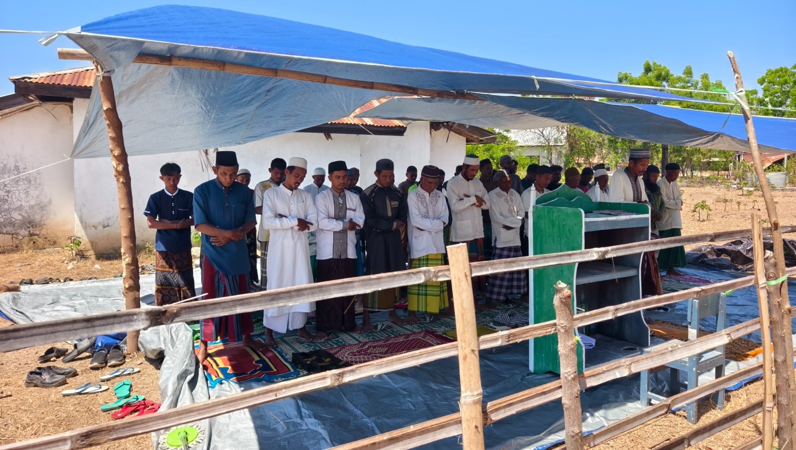 Suasana shalat berjamaah warga Waekokak di mushola darurat dengan mimbar kayu sederhana di bawah naungan atap terpal biru.